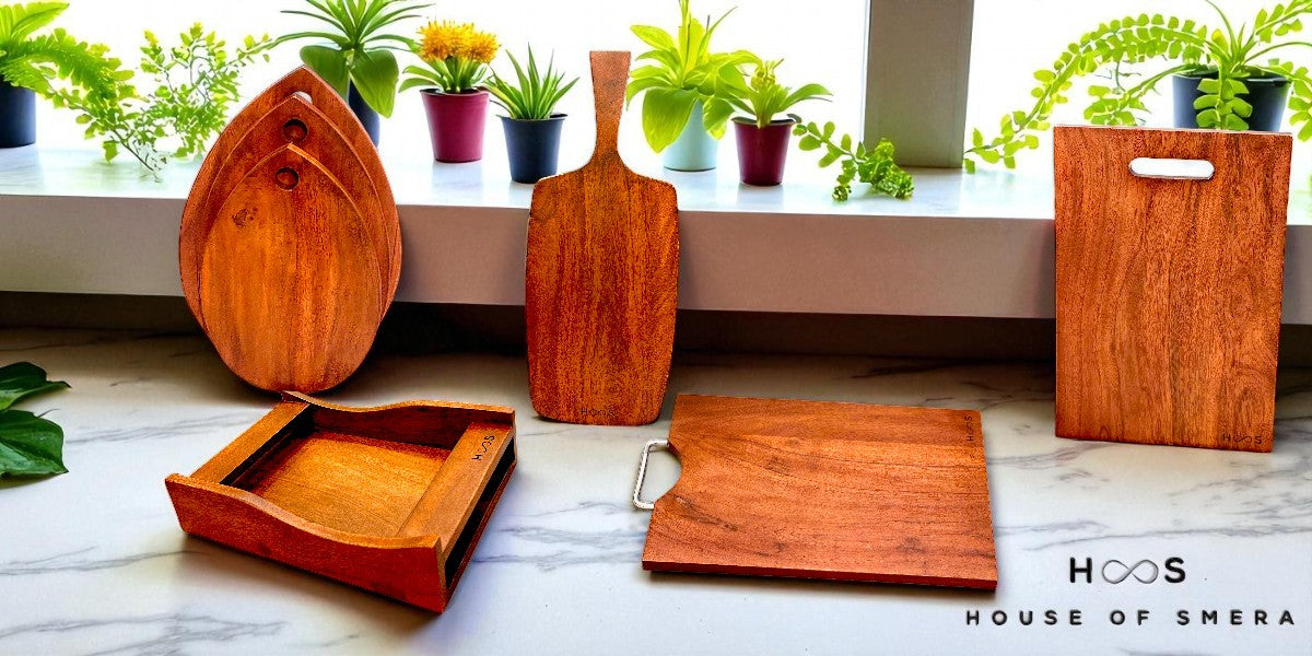 Wooden cutting boards and trays on a marble surface with plants in the background, featuring the brand 'House of Shera'.