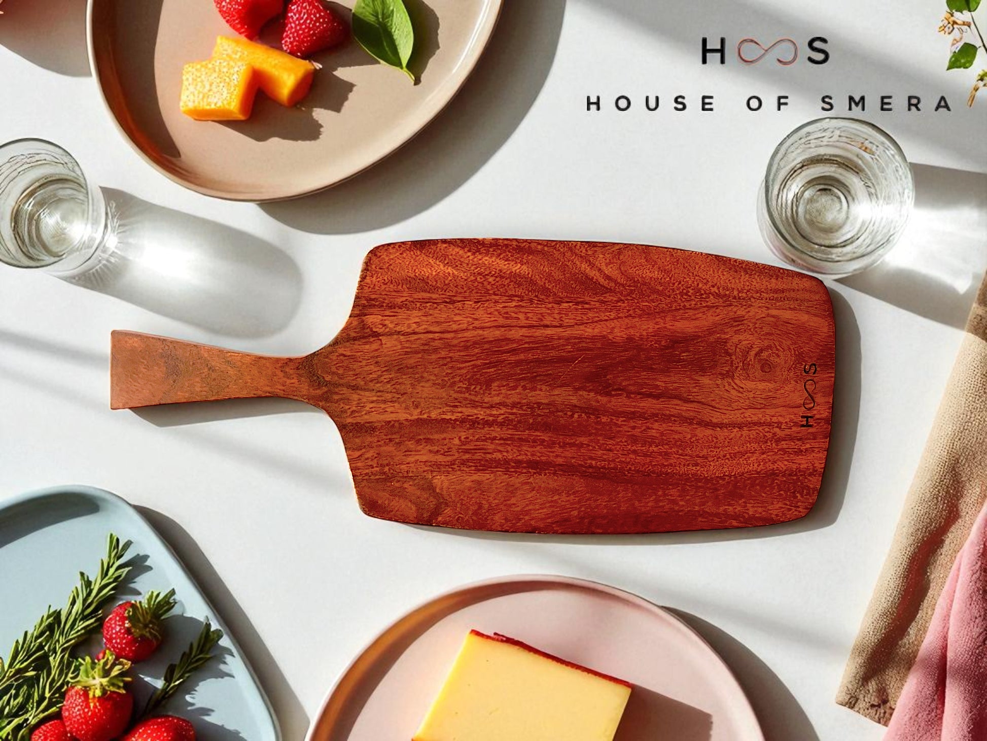 Wooden cutting board on a table with plates of fruit and cheese, and 'House of Smera' branding.